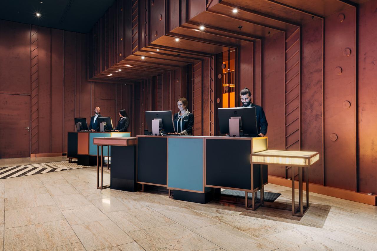 Pullman Berlin Schweizerhof Hotel Ngoại thất bức ảnh The image shows a modern hotel lobby reception area. There are several reception desks made of dark materials with illuminated accents. Behind the desks, three people are standing, likely staff members, engaged with computer screens. The lobby featur